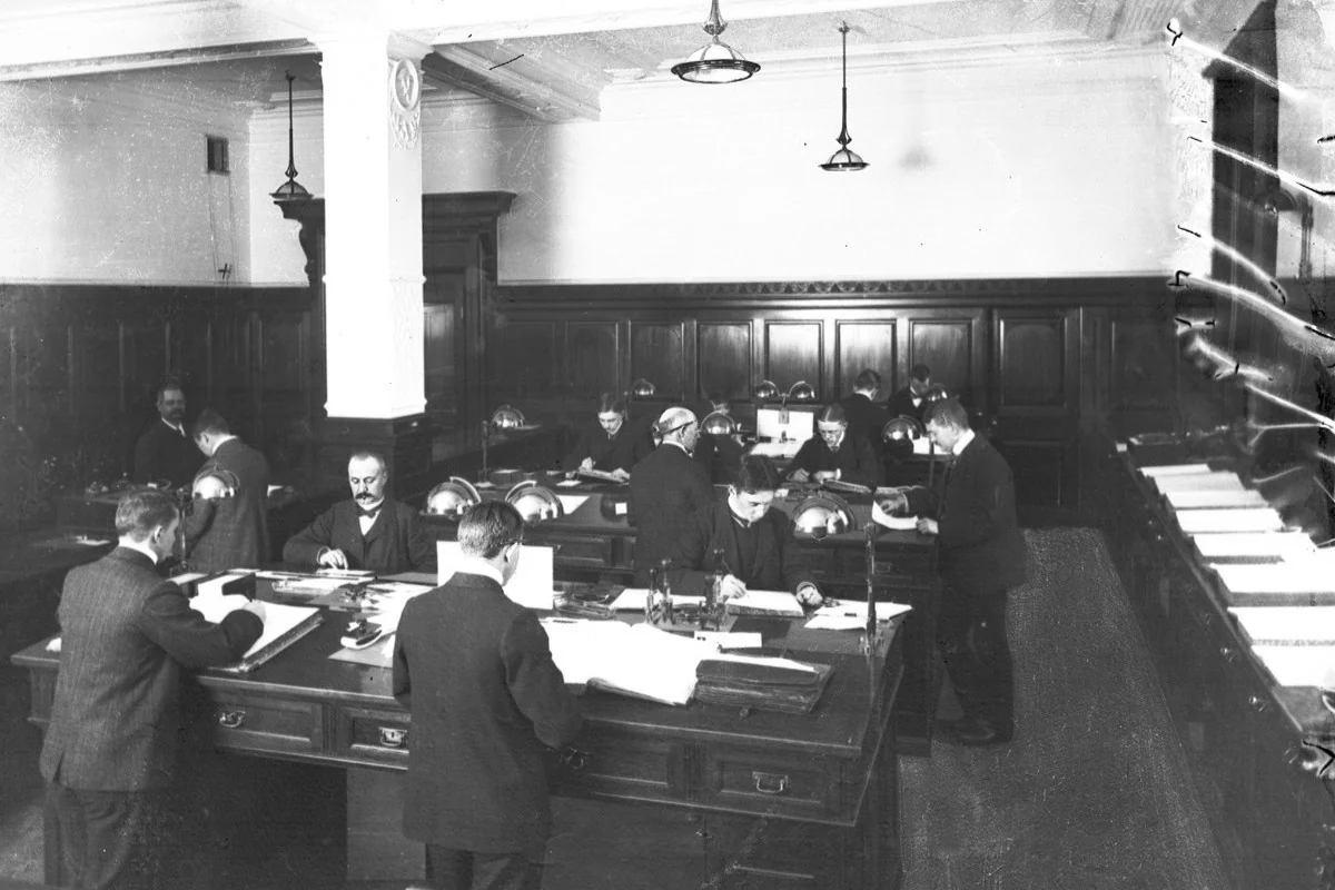 Sydjylland 1906: Mød personalet i Vejle Bank. Ved forreste pult ses fuldmægtig C.A. Christensen og assistent C. Jensen, begge med ryggen til. Overfor ses assistent B.M. Møller og assistent Konradsen. Ved mellemste pult ses - med ryggen til - direktør Jacob Petersen og overfor assistent Loye, assistent Max Christiansen samt bogholder Aage Petersen. Ved tredje pult, assistent Wentzel og overfor assistent C. Simonsen. Ved skranken kasserer Emil Rasmussen. (Foto: Vejle Stadsarkiv)