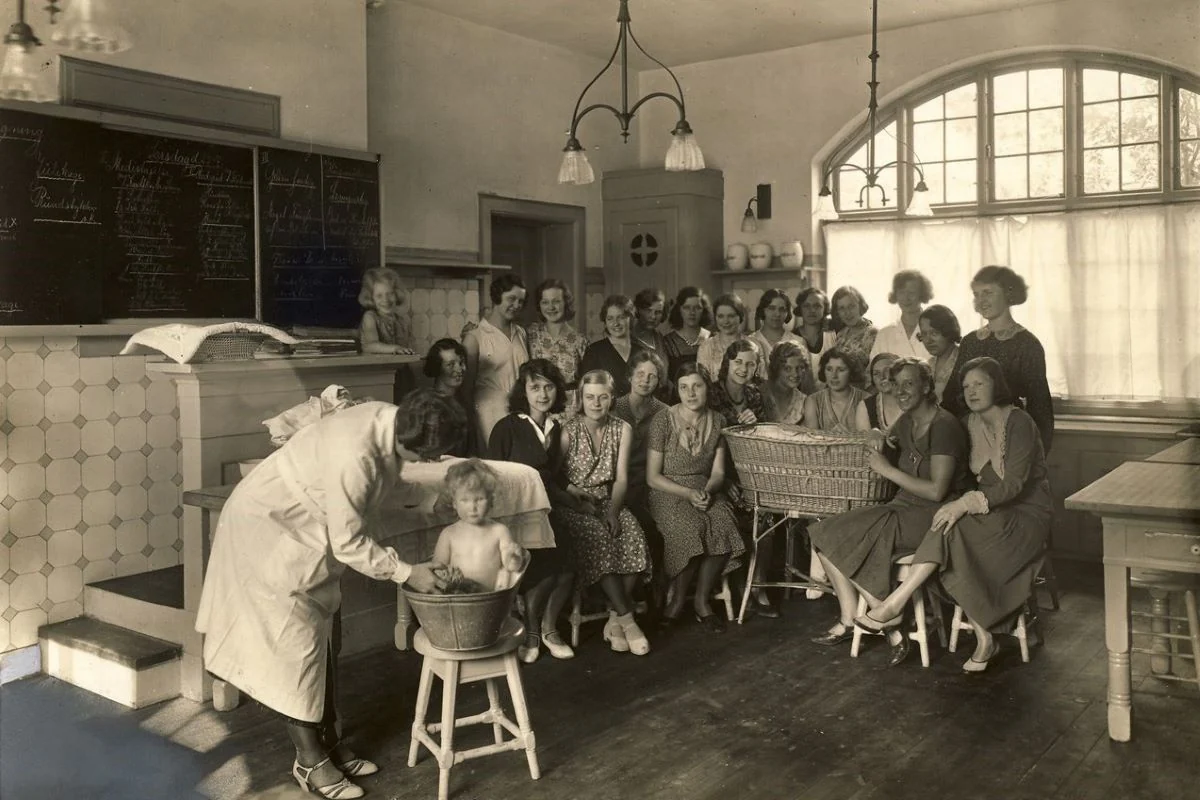 <b>Nordsjælland 1908: Holte Husholdningsskole. Spædbørnspleje.</b> Pigerne skulle lære at holde hus - og pleje deres babyer. Her scene fra undervisning i klasselokalet. Pigen ved katederet fjerner bordet i venstre hjørne. Huset opført i 1908. Skolen åbnede i 1909. (Foto: Historisk Arkiv for Rudersdal Kommune)