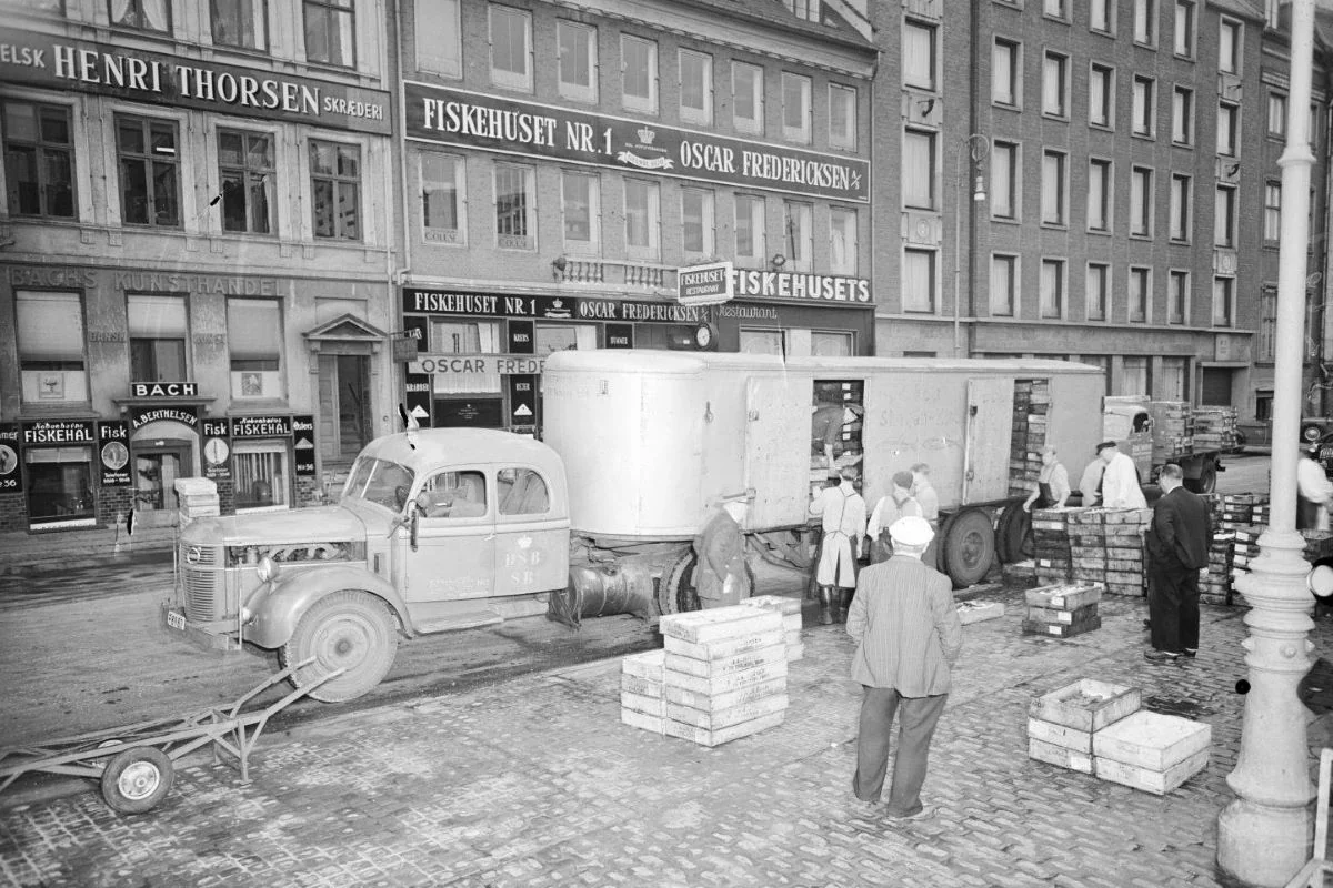 <b>København 1951: Der læsses frisk fisk af i Indre By.</b> En tidlig morgen - der gøres klar til dagens handel. Skagensbanen og Danske Statsbaners fragtrute for fisk <i>Skagen-Grenå-København-Hundested</i> udlæsser gods ved fiskemarkedet på Gammel Strand i København. I baggrunden ses nogle af datidens fiskerestauranter i gaden. Billedet er taget før solopgang kl. 05.10, og der er derfor benyttet blitz ved optagelsen. (Foto: Danmarks Jernbanemuseum)