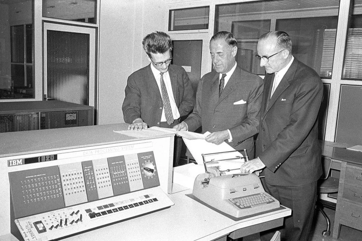 <b>Fyn 1961: Ny elektronhjerne til maskinfabrikken Thrige.</b> Scene fra computerens barndom: En tekniker i selskab med direktør Aage Lommer og underdirektør Henry Vinding ved det nye edb-anlæg, en IBM 1620, der var kommet på markedet i 1959. Det var den første computer, der ansås som stabil nok til at styre produktionsprocesser til brug i industrisamfundet. (Foto: Odense Stadsarkiv)