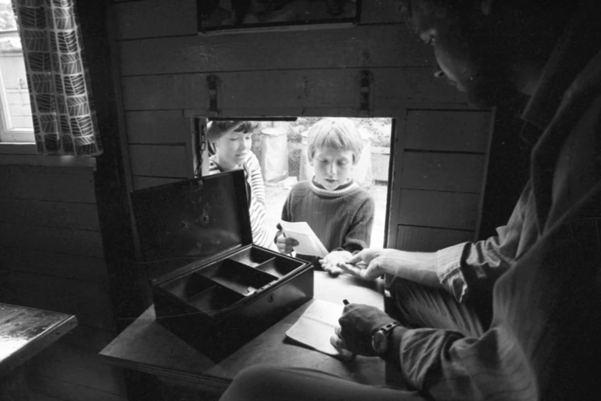 Østfyn 1969: Børnene går i Kolonibanken. Københavns Lærerforening drev i juli 1969 feriekolonien Knuds Minde i Nyborg, hvor 54 drenge kunne få sol på kroppen i sommerens løb. Drengenes lommepenge blev ved ankomsten anbragt i Kolonibanken, der stort set kun var en pengekasse, hvorfra de kunne hæve penge, som oftest til køb af is, sodavand eller lignende. På billedet har en af de unge sparere, Stig, stillet sig op ved kassererlugen i Kolonibanken for at få udbetalt 50 øre. De skulle bruges til et frimærke, der skulle på et brev til familien. (Foto: Odense Stadsarkiv)