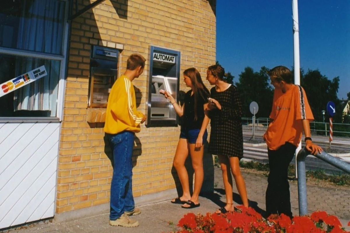 <b>Østjylland 1997: Byens første hæveautomat.</b> Nu kan man hæve penge uden at skulle i banken. Her benyttes Sparekassen Kronjyllands nye hæveautomat af Gitte Kjærsgaard fra Aarslev og Anne Bundgaard Kristensen fra Hørning, mens to ungersvende ser på. Den 8. august 1997 i Assentoft. (Foto: Aalborg Stadsarkiv)